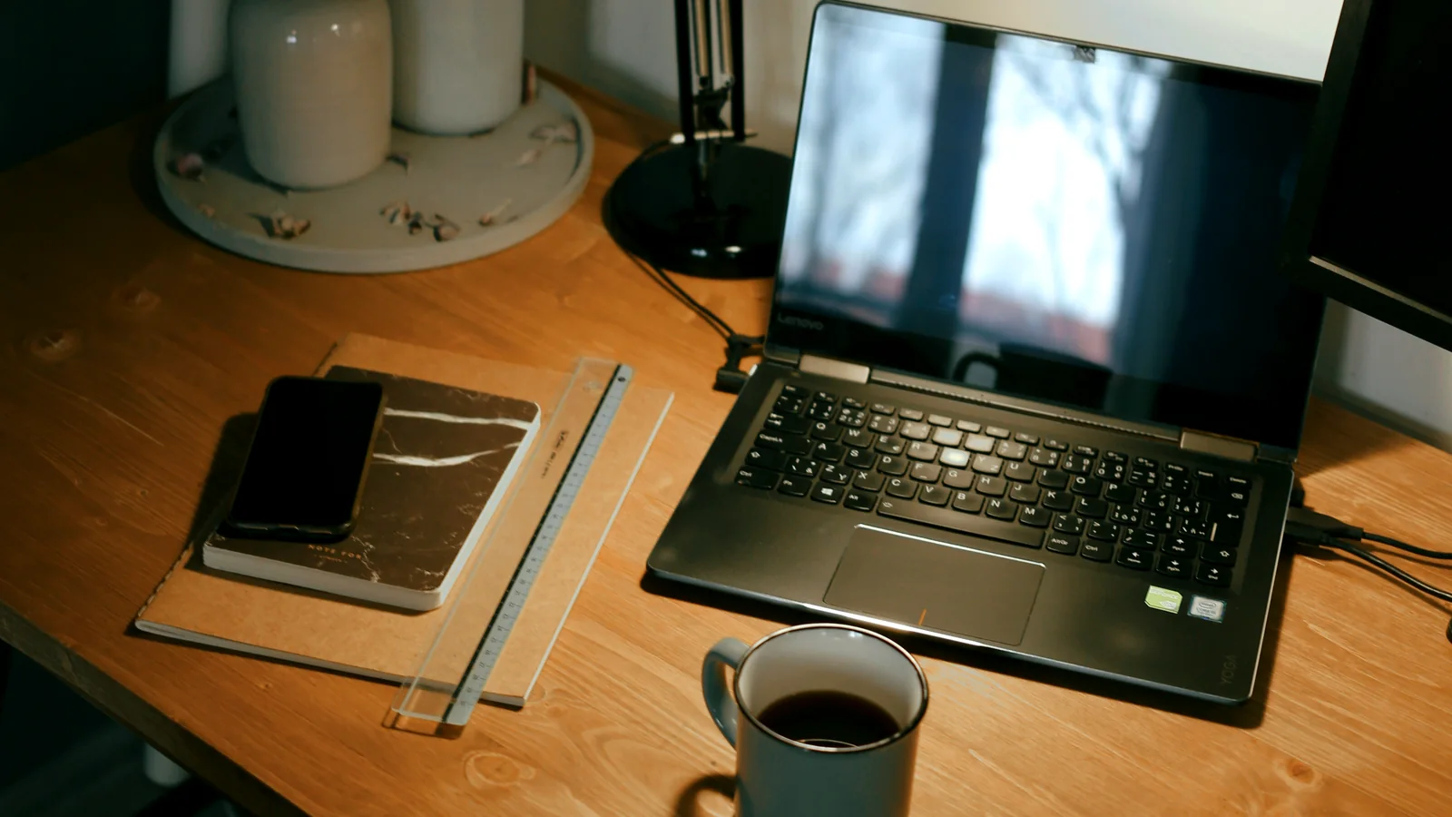 Mesa de trabalho realista com notebook, celular virado para baixo, caderno, régua e xícara em luz quente.